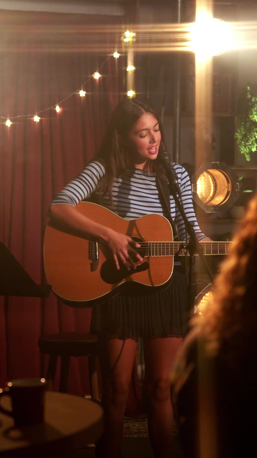 Strings Attached — Sadie singing and playing guitar on a small stage with string lights